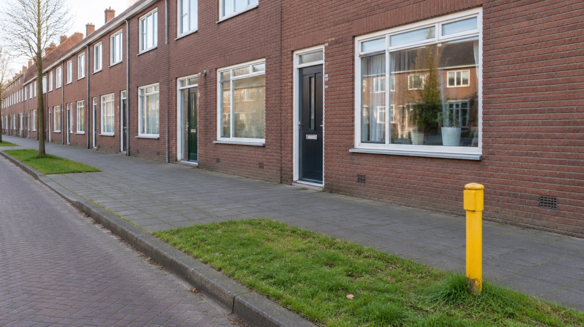 Kleine gele markeerpaal van een gasleiding in een grasberm langs een voetpad, met bakstenen rijtjeswoningen op de achtergrond in het ochtendlicht