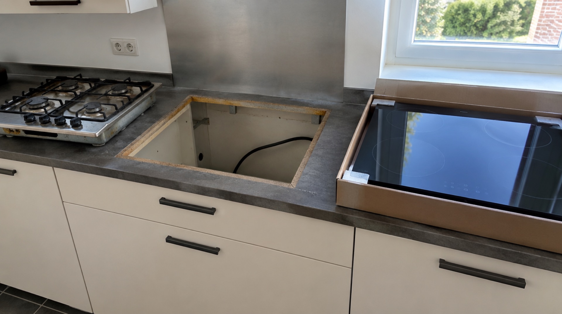 Keuken tijdens een verbouwing, met een lege uitsparing in het aanrecht, ernaast een uitgebouwde oude gaskookplaat en een kartonnen doos met een nieuwe inductiekookplaat in het daglicht