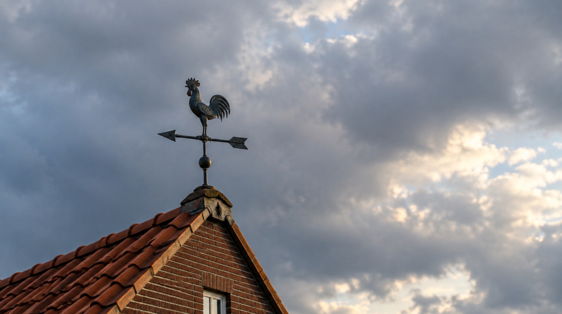 Windvaan in de vorm van een haan op de nok van een pannendak tegen een wisselende, bewolkte lucht