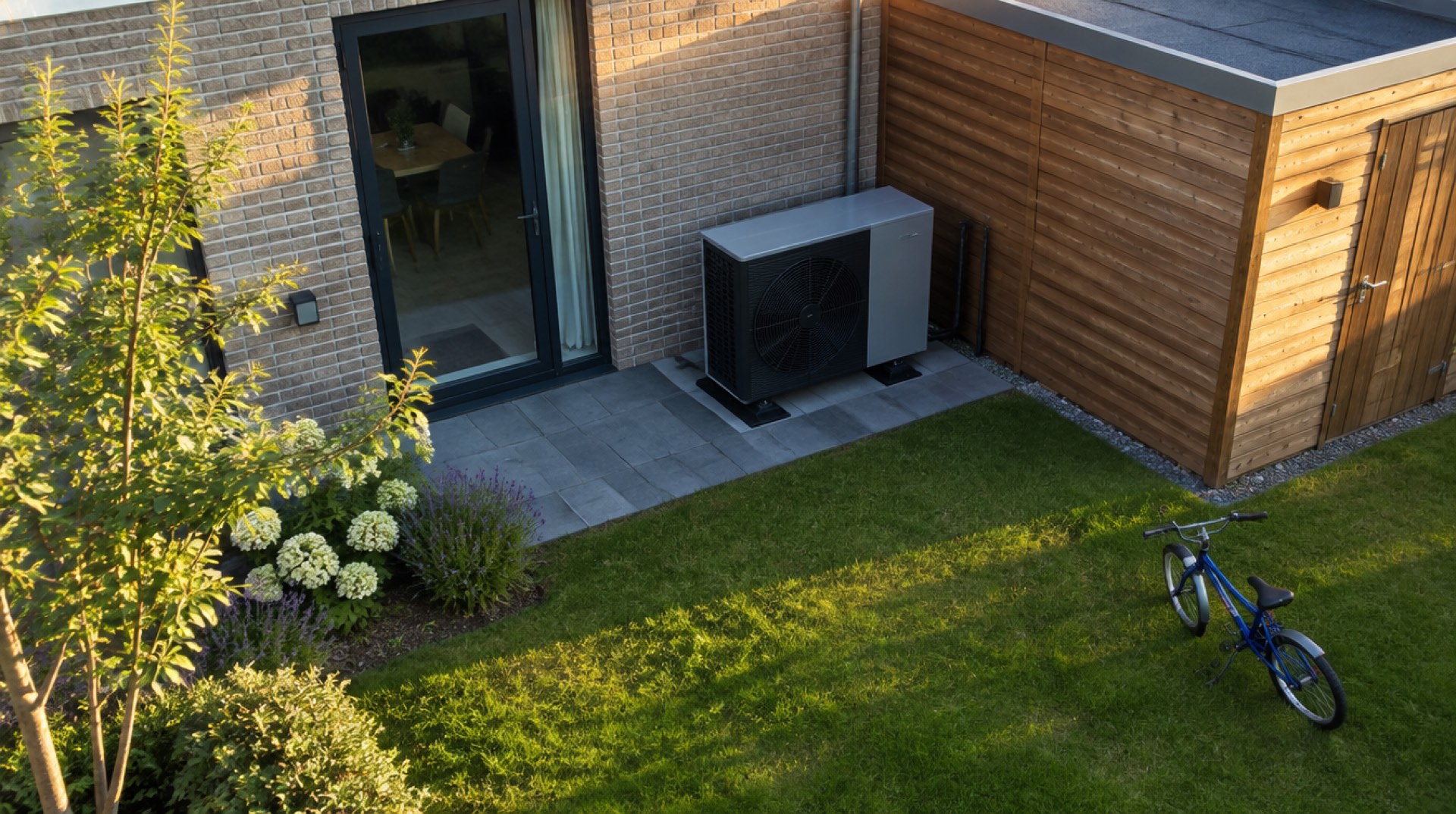 Warmtepomp buitenunit bij een nieuwbouwwoning en houten schuur, met een fiets op het gras